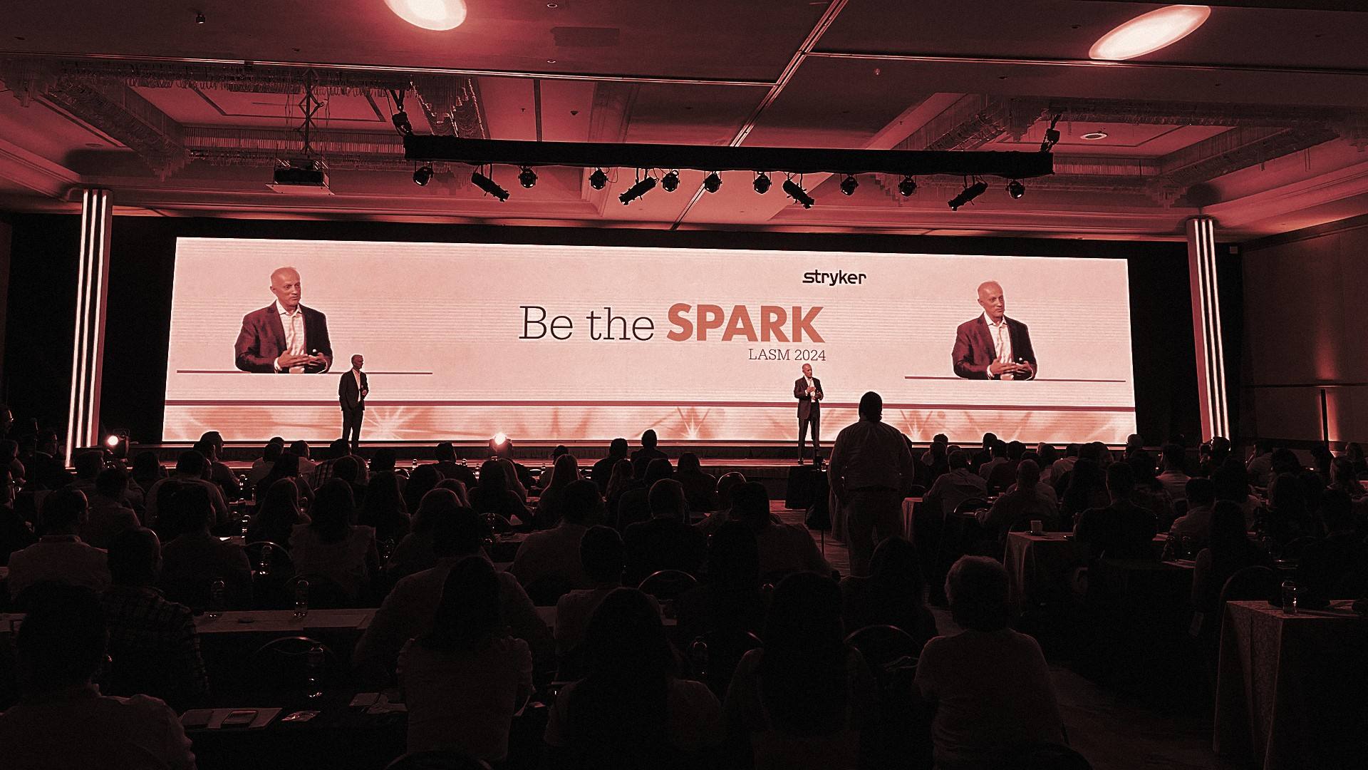 General session of Be the Spark: two speakers on stage in front of the main screen, the audience in silhouette