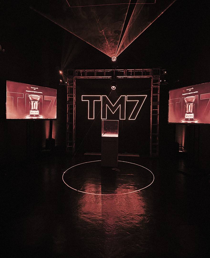 Escenario del lanzamiento TM7 en penumbra, con la vitrina al centro y las pantallas laterales